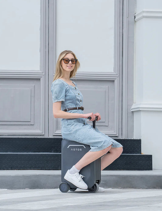 A stylish blonde woman in a denim dress and sunglasses sits comfortably on a dark grey AOTOS L2 suitcase, which has large, smooth-rolling wheels.