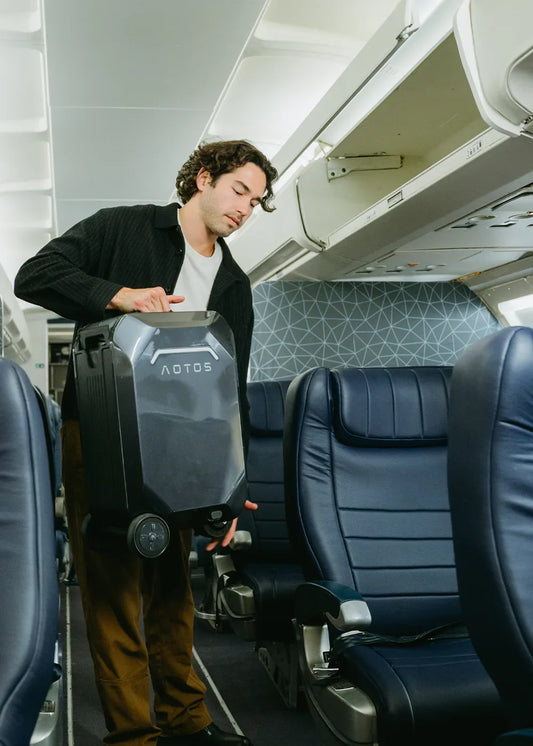 Traveler lifting AOTOS smart rideable luggage into airplane cabin aisle—carry-on compliant electric suitcase for effortless in-flight mobility.