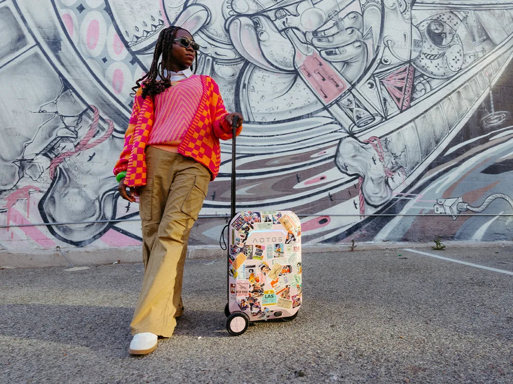 Trendsetting traveler pulling a sticker-covered AOTOS smart rideable suitcase past a vibrant street-art mural, highlighting personalized carry-on, ride-on mobility.