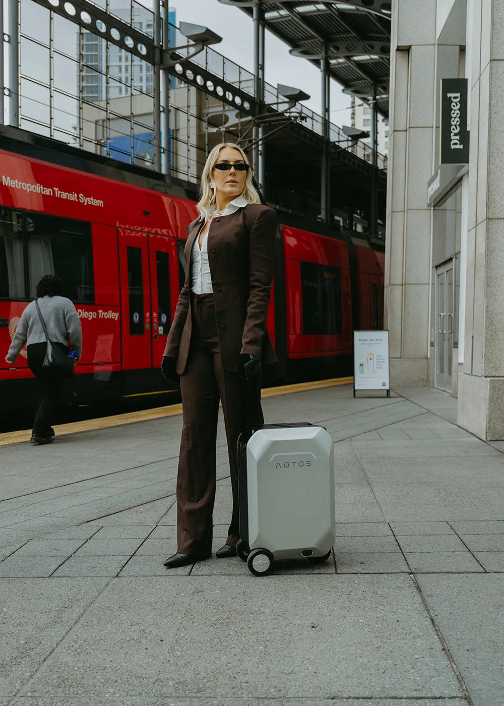A stylish woman in a brown suit stands on a train platform next to her light gray Aotos L2 smart rideable suitcase, with a red trolley in the background.
