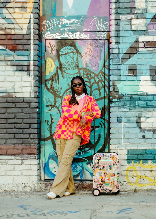 A person in a vibrant pink and orange checkered cardigan and khaki pants stands casually against a graffiti-covered wall, next to an AOTOS L2 smart rideable suitcase adorned with colorful stickers.