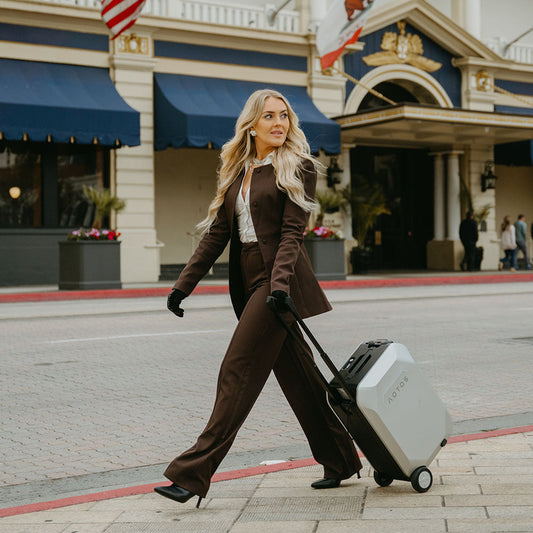 Business traveler pulling an AOTOS ride-on smart suitcase past a city hotel entrance.