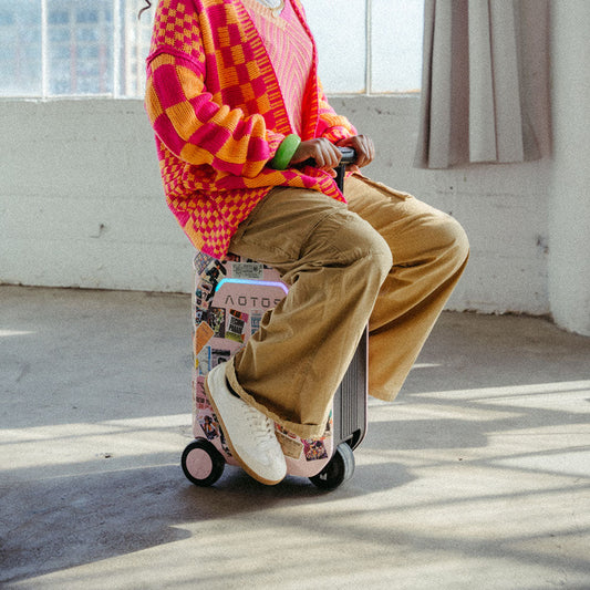 Person riding an AOTOS smart rideable suitcase decorated with travel stickers, sitting indoors in natural sunlight.