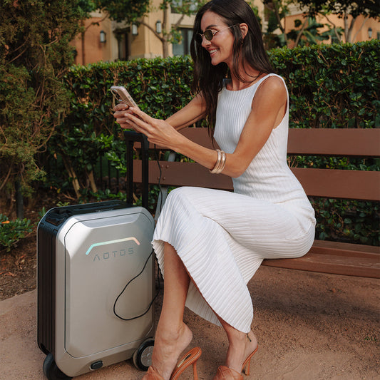 Woman sitting on a bench charging her phone with a sleek silver AOTOS smart luggage, wearing a white dress and sunglasses outdoors.