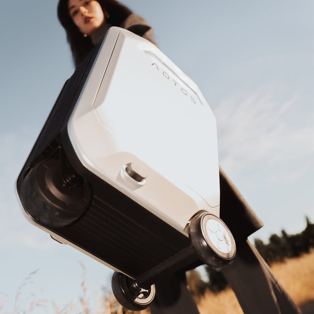 Close-up low-angle view of a traveler holding the Aotos L2 smart suitcase with durable spinner wheels, photographed outdoors under a clear sky.