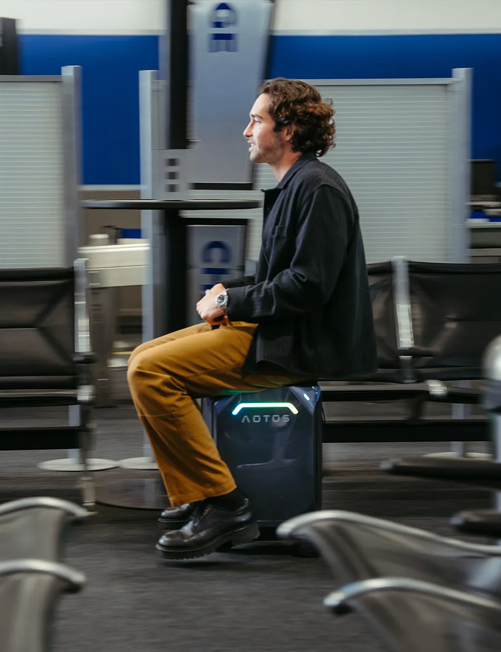 Man effortlessly riding an AOTOS L2 motorized smart suitcase through an airport terminal for seated, hands-free mobility